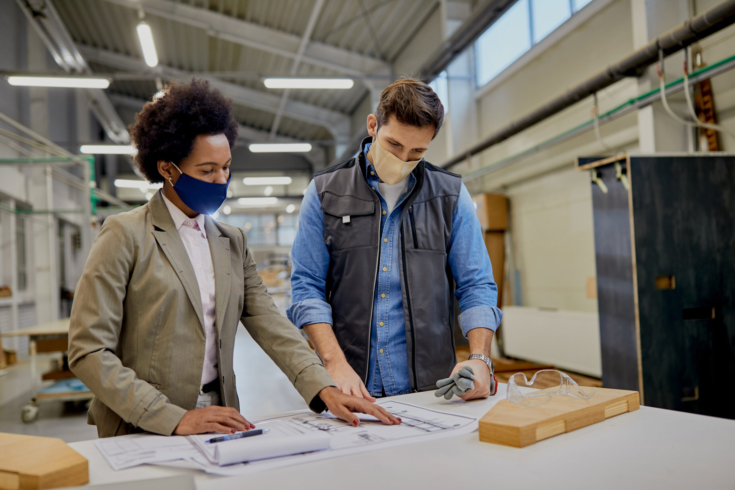 African American female manager and carpenter wearing face masks while analyzing design plans at production facility.